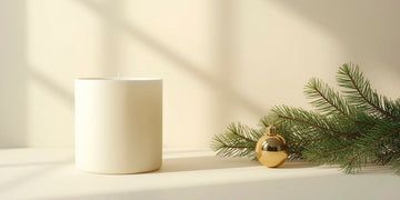 Simple Christmas decor: cream candle, pine branch, and gold bauble on a plain beige surface with soft shadows.