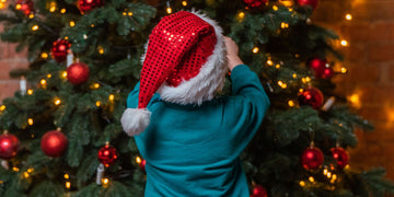 A child in a red sequined Santa hat reaches up to hang an ornament on a Christmas tree.